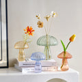 Quatre vases champignon en verre coloré, ambré, vert, bleu et rosé, posés sur des livres avec des fleurs