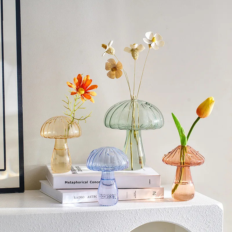 Quatre vases champignon en verre coloré, ambré, vert, bleu et rosé, posés sur des livres avec des fleurs